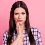 Brunette woman in flannel against a pink wall cringes in pain due to tooth decay and touches her cheek
