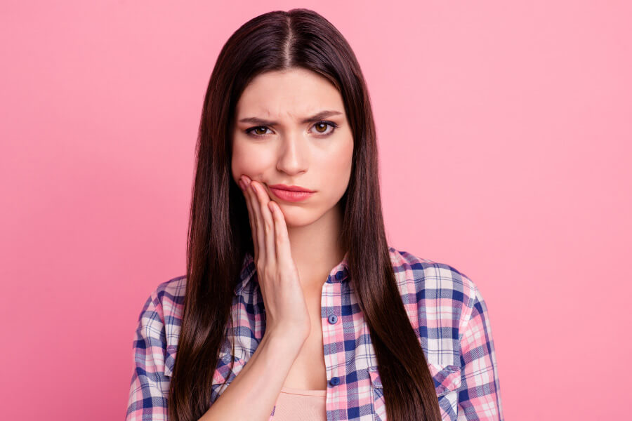 Brunette woman in flannel against a pink wall cringes in pain due to tooth decay and touches her cheek in Bellevue