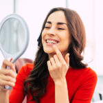 Brunette woman in a red blouse smiles at herself in a handheld mirror after professional teeth whitening