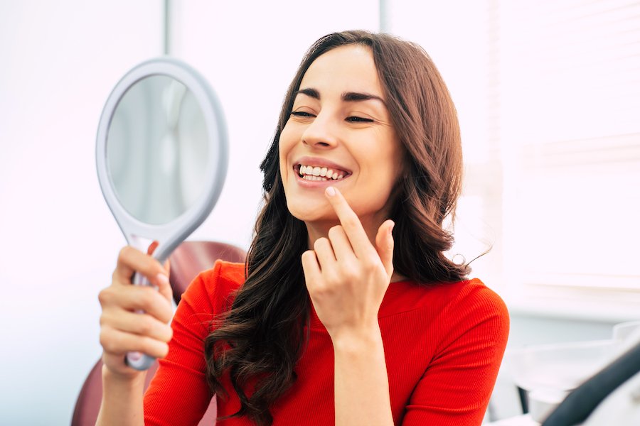 Brunette woman in a red blouse smiles at herself in a handheld mirror after professional teeth whitening in Bellevue