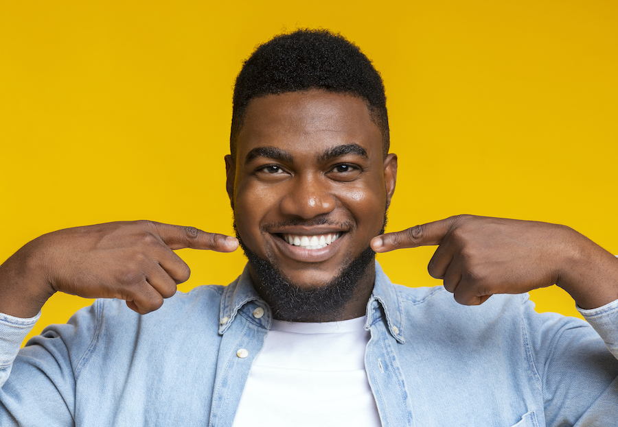 A Black man smiles and points to his teeth while standing against a yellow background