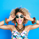 Black woman smiles while wearing sunglasses and chunky jewelry in front of a blue background