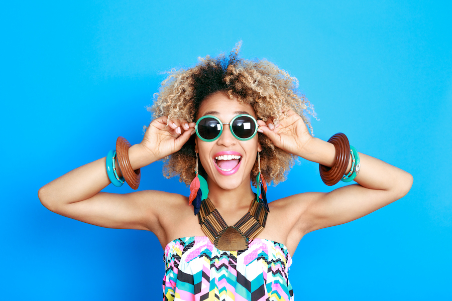 Black woman smiles while wearing sunglasses and chunky jewelry in front of a blue background
